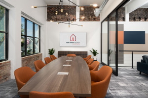 HQ on Main teleconference room with ten orange swivel chairs and wood table with integrated technology and modern ceiling pendant lighting television with Red Door Agency logo